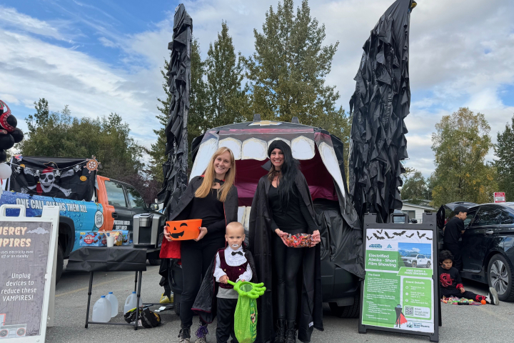 Two women and a child, all dressed as vampires, stand in front on an electric vehicle's frunk that is decorated to look like a vampire bat.