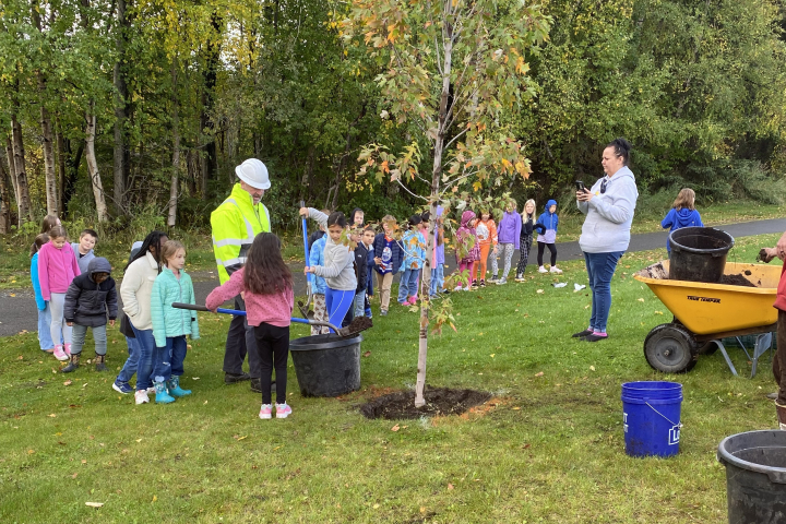 Children line up to shovel dirt into the hole where a new tree is being planted. A man in a reflective coat and white hard hat watches.