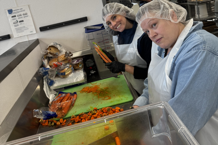 Two volunteers in aprons and hairnets peel and chop carrots.
