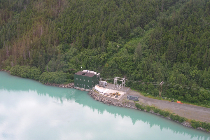 The Cooper Lake dam power house sits on the water's edge with with a forest background. Powerlines extend out from the building.