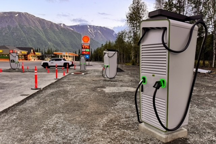 A white electric vehicle charger sits behind cones in a parking lot. A store and gas station are in the background.