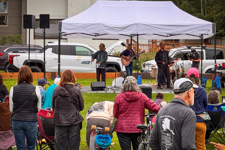A crowd watches musicians perform under a white tent at an outdoor event. A Chugach Electric RV truck is parked behind them.