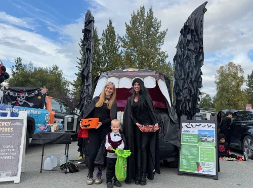 Two women and a child, all dressed as vampires, stand in front on an electric vehicle's frunk that is decorated to look like a vampire bat.