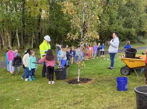 Children line up to shovel dirt into the hole where a new tree is being planted. A man in a reflective coat and white hard hat watches.