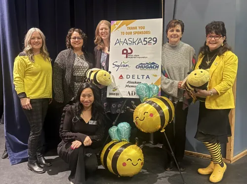 2026 Alaska Spelling Bee volunteers gather around a sponsorship poster.