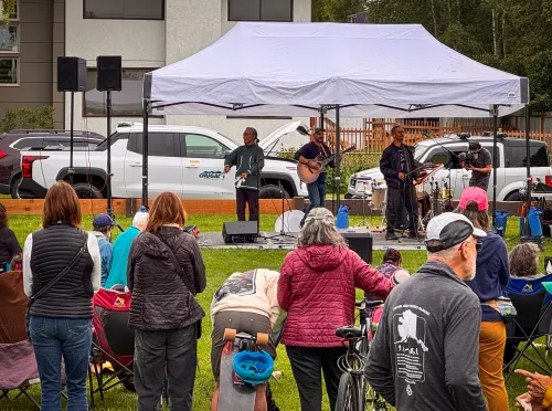 A crowd watches musicians perform under a white tent at an outdoor event. A Chugach Electric RV truck is parked behind them.