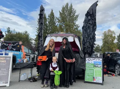 Two women and a child, all dressed as vampires, stand in front on an electric vehicle's frunk that is decorated to look like a vampire bat.