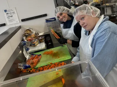 Two volunteers in aprons and hairnets peel and chop carrots.