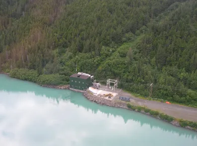 The Cooper Lake dam power house sits on the water's edge with with a forest background. Powerlines extend out from the building.