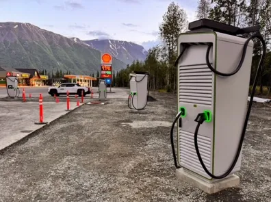 A white electric vehicle charger sits behind cones in a parking lot. A store and gas station are in the background.
