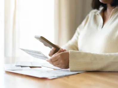 A person in a white sweater sits at a table with bills on it, holding a bill in one hand and a cell phone in the other.