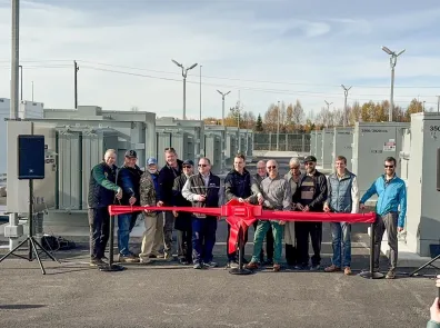 Group poses with scissors on a big red ribbon with a bow. BESS batteries are in the background.