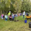 Children line up to shovel dirt into the hole where a new tree is being planted. A man in a reflective coat and white hard hat watches.