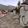 A white electric vehicle charger sits behind cones in a parking lot. A store and gas station are in the background.