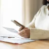 A person in a white sweater sits at a table with bills on it, holding a bill in one hand and a cell phone in the other.