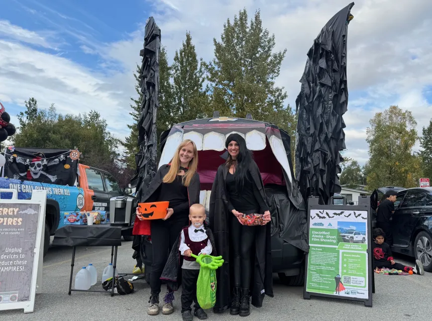 Two women and a child, all dressed as vampires, stand in front on an electric vehicle's frunk that is decorated to look like a vampire bat.