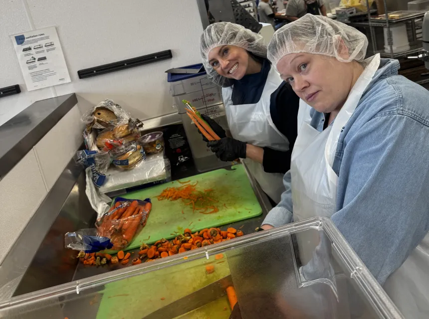 Two volunteers in aprons and hairnets peel and chop carrots.