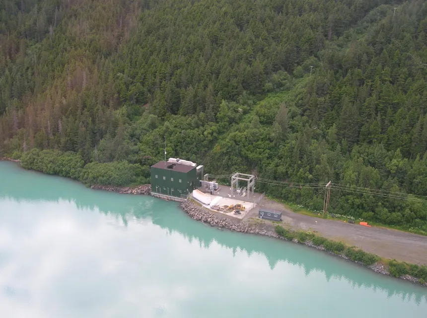 The Cooper Lake dam power house sits on the water's edge with with a forest background. Powerlines extend out from the building.
