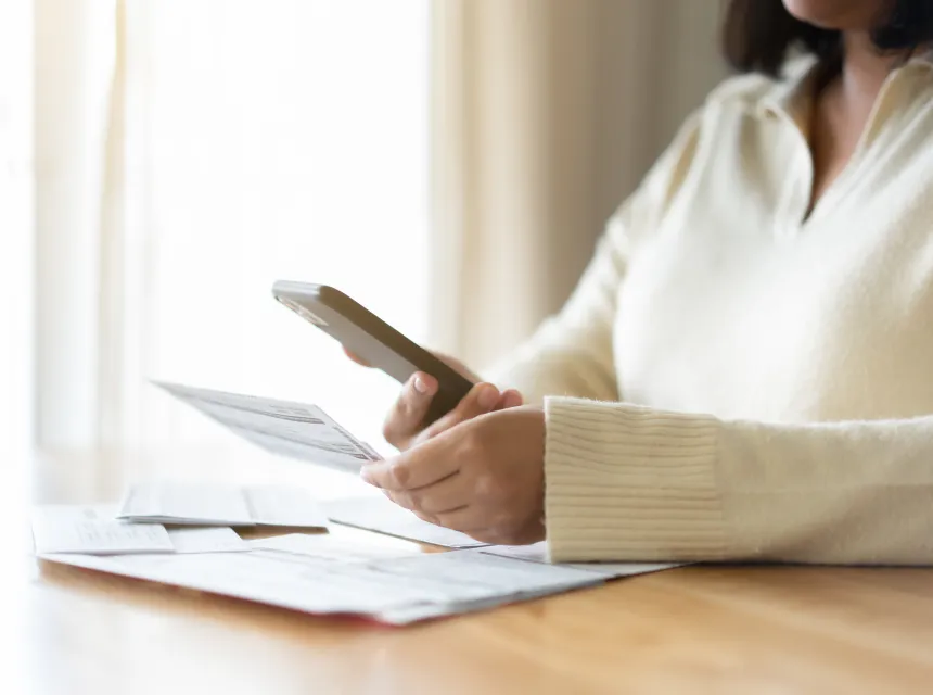 A person in a white sweater sits at a table with bills on it, holding a bill in one hand and a cell phone in the other.